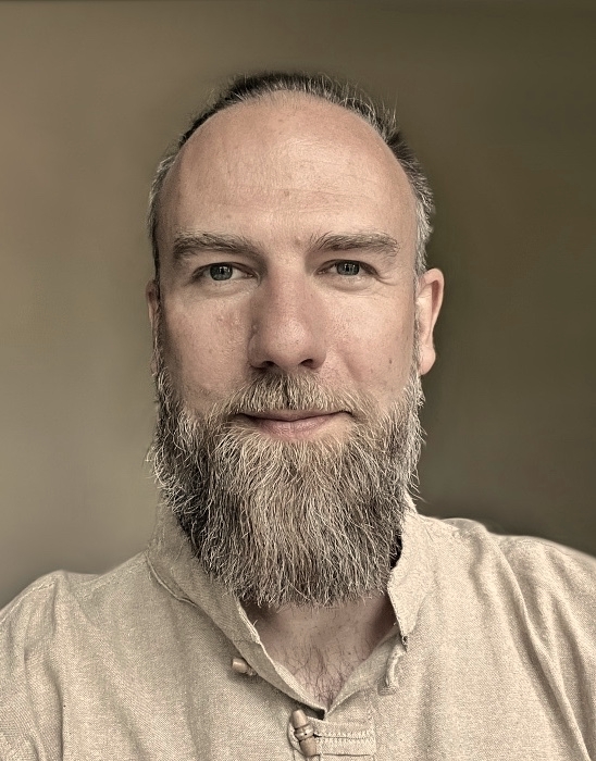 Portret psychoterapeuty, Michała Lehr-Kowalskiego -lekko uśmiechniętego mężczyzny z jasną brodą w lnianej koszuli w beżowym kolorze, o wschodnim kroju.